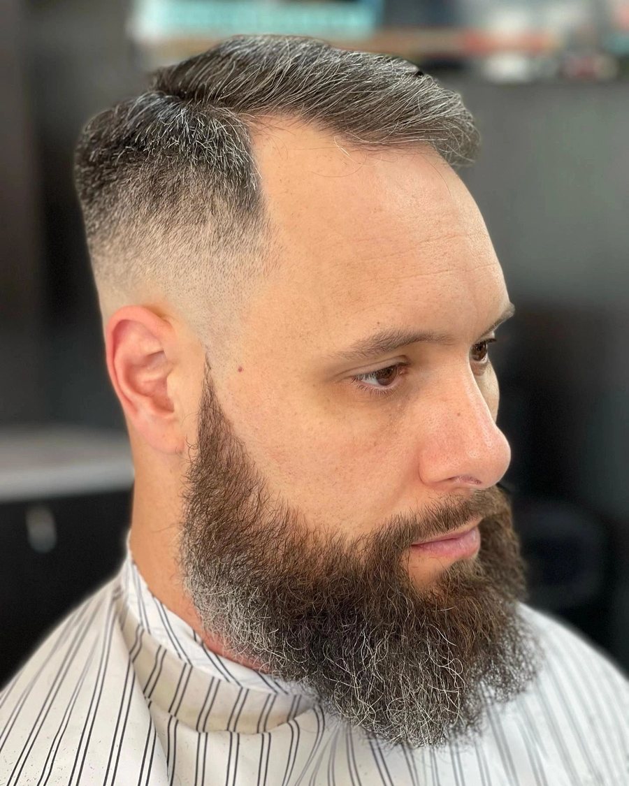 Man facing the camera with a full beard shaped to a high fade.